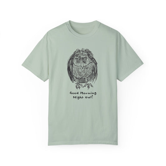 Alt text: Light green or sage colored t-shirt featuring an illustration of a tired-looking owl with messy feathers, created from a linoleum block print. The owl stands with drooping eyes, and below it, the text reads: “Good Morning Night Owl!