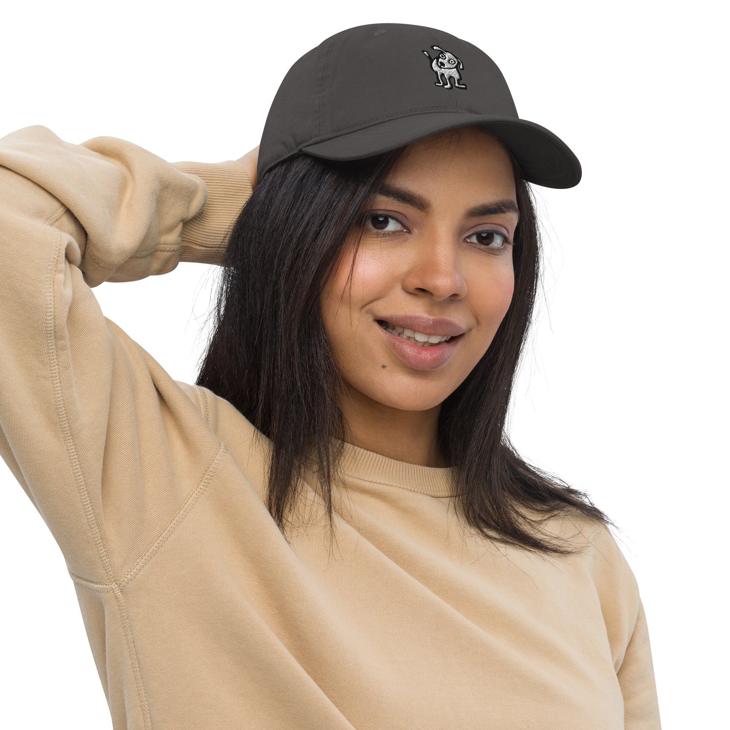 Woman wearing a charcoal color baseball cap with an embroidered cartoon-style dog logo on the front, smiling and posing with one hand resting on the back of her head; she is dressed in a beige sweatshirt and has long, dark hair.