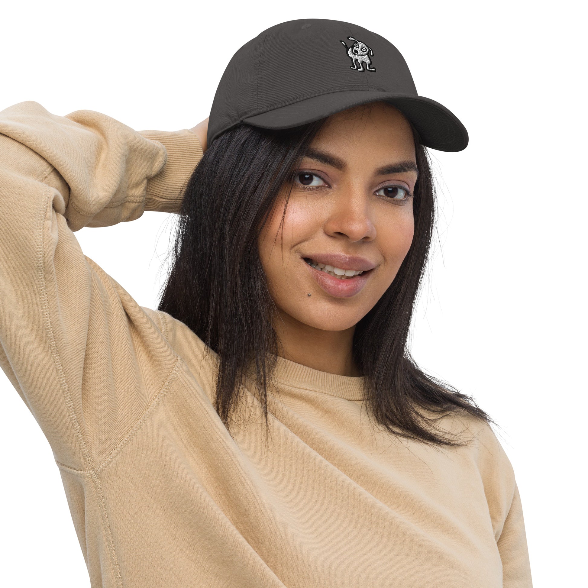 Woman wearing a charcoal color baseball cap with an embroidered cartoon-style dog logo on the front, smiling and posing with one hand resting on the back of her head; she is dressed in a beige sweatshirt and has long, dark hair.