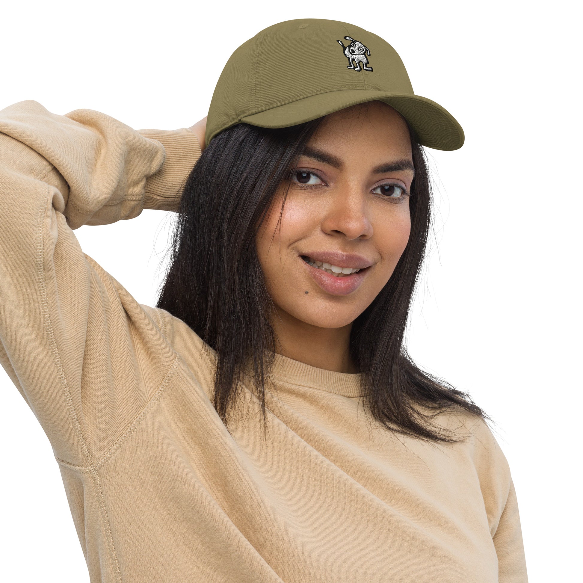 Woman wearing a jungle green baseball cap with an embroidered cartoon-style dog logo on the front, smiling and posing with one hand resting on the back of her head; she is dressed in a beige sweatshirt and has long, dark hair.