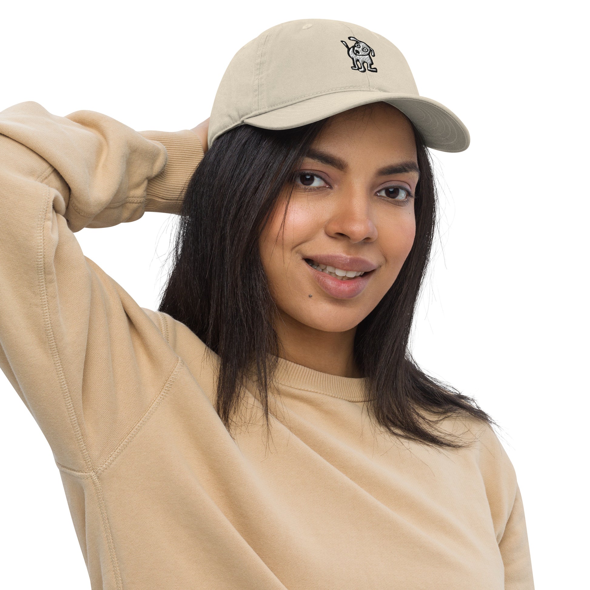 Woman wearing a off-white baseball cap with an embroidered cartoon-style dog logo on the front, smiling and posing with one hand resting on the back of her head; she is dressed in a beige sweatshirt and has long, dark hair.