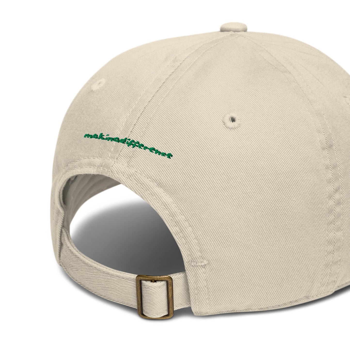 Rear view of a off-white baseball cap featuring an adjustable brass buckle strap and small green embroidered text that reads "makinadifference" above the opening.