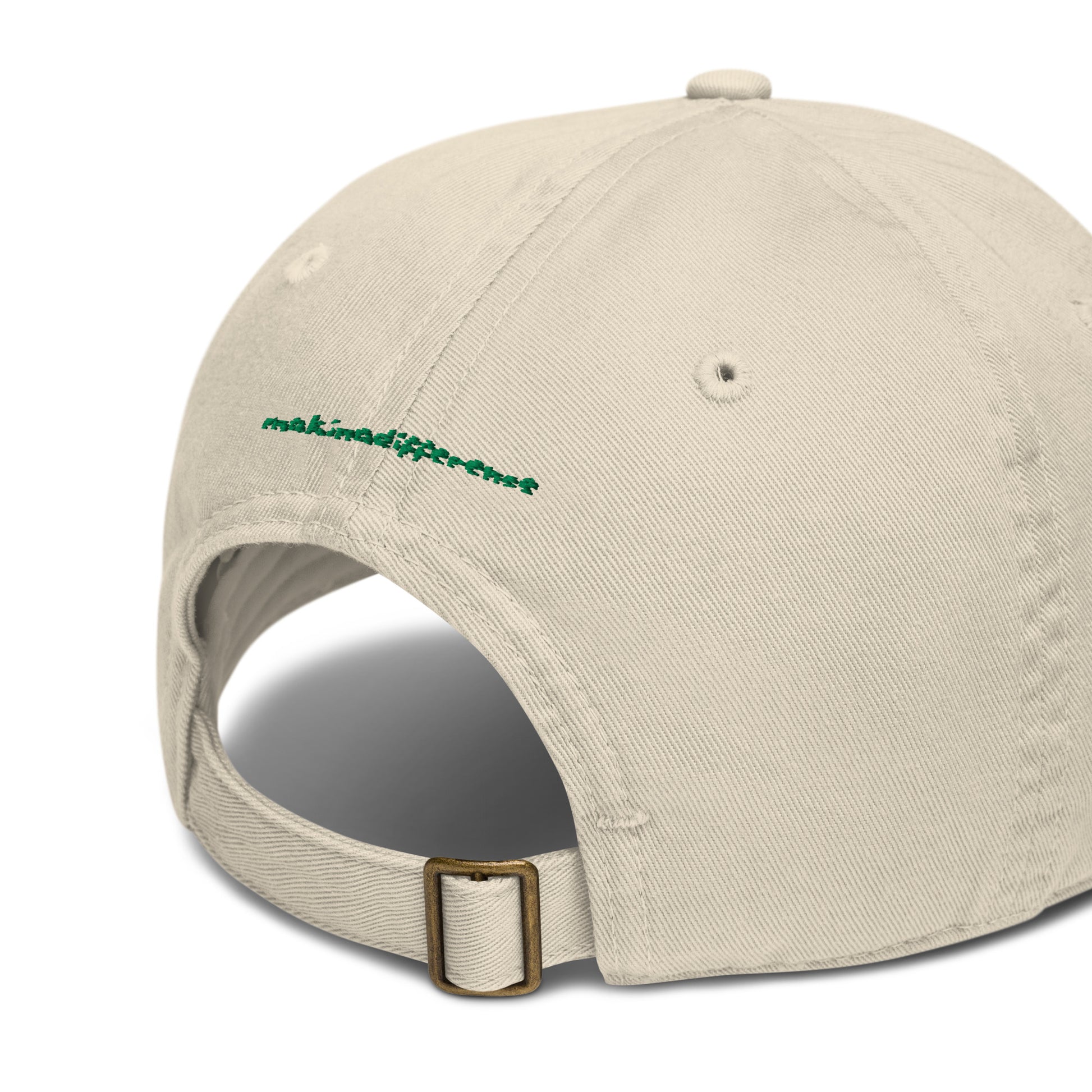 Rear view of a off-white baseball cap featuring an adjustable brass buckle strap and small green embroidered text that reads "makinadifference" above the opening.