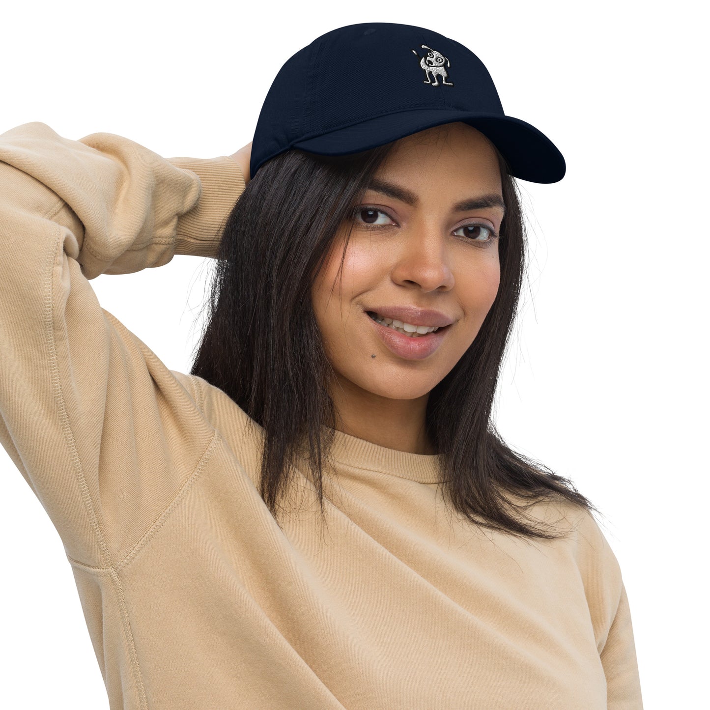 Woman wearing a navy blue baseball cap with an embroidered cartoon-style dog logo on the front, smiling and posing with one hand resting on the back of her head; she is dressed in a beige sweatshirt and has long, dark hair.