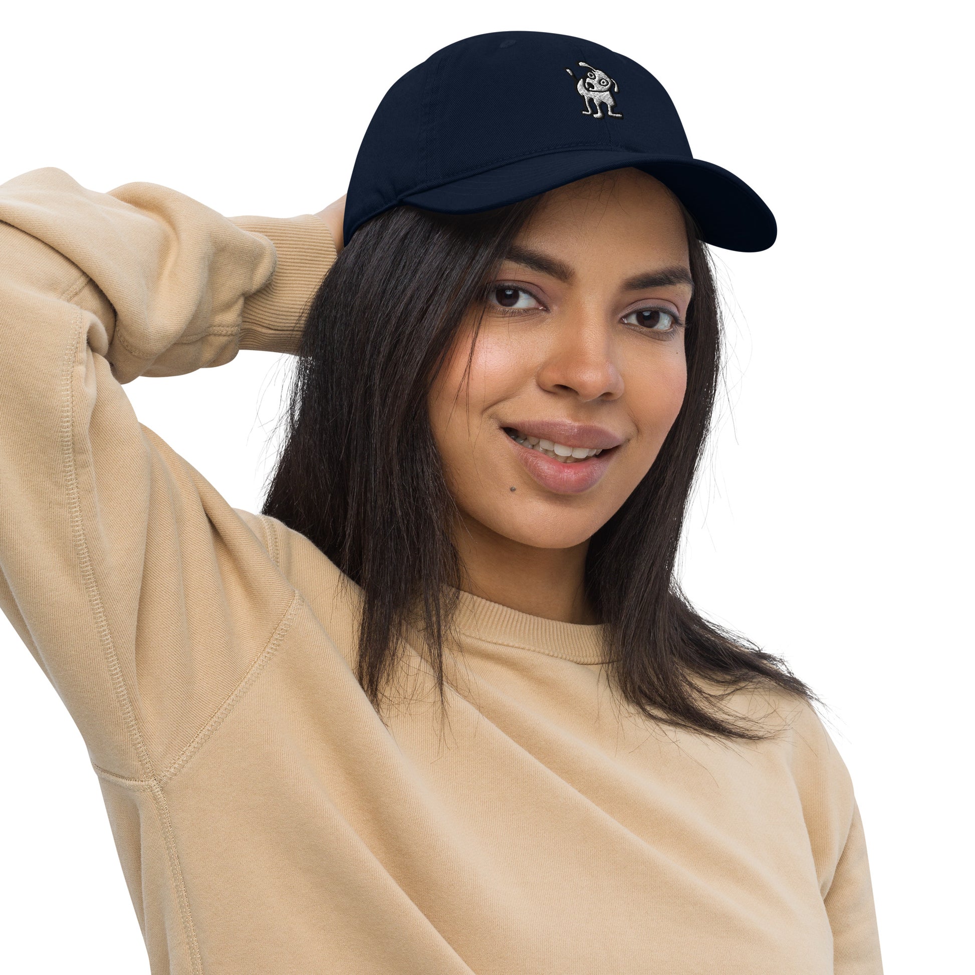 Woman wearing a navy blue baseball cap with an embroidered cartoon-style dog logo on the front, smiling and posing with one hand resting on the back of her head; she is dressed in a beige sweatshirt and has long, dark hair.