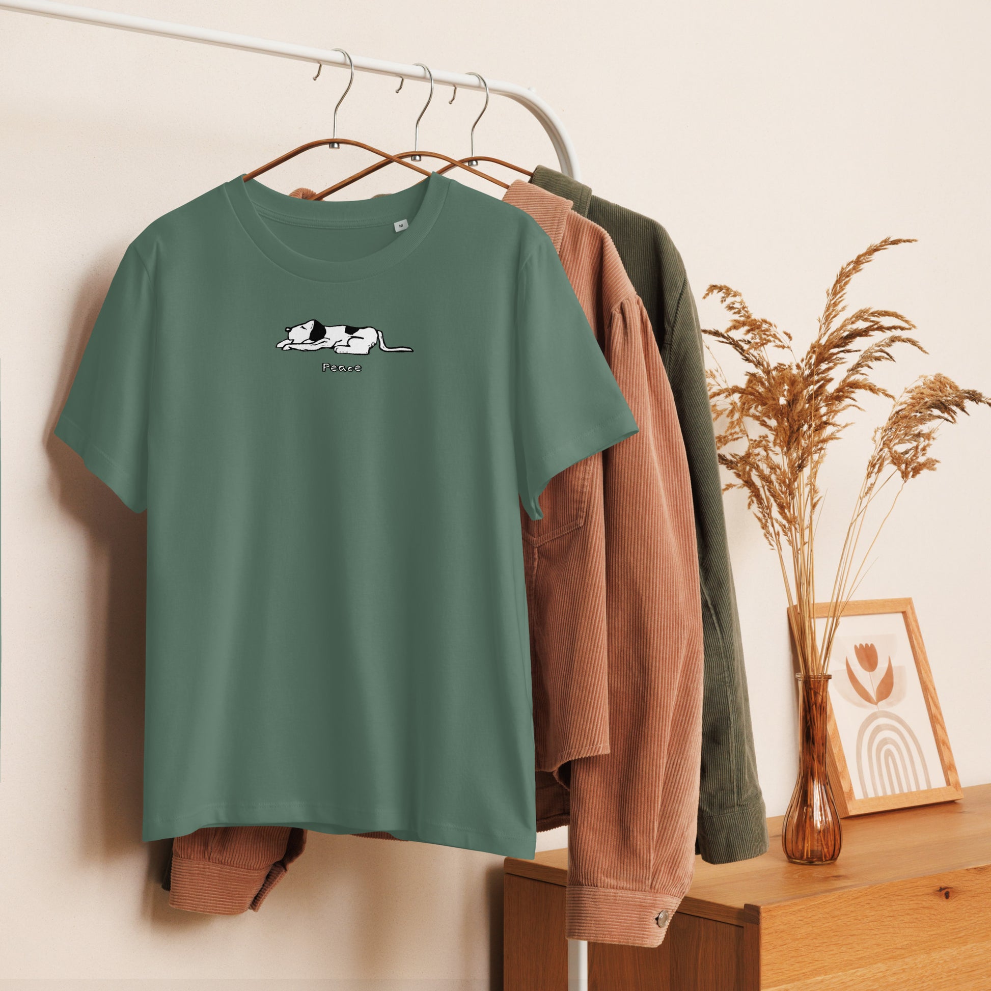 Green unisex t-shirt hanging on a clothing rack, featuring a black and white cartoon-style dog lying down with the word “Peace” written below. Surrounding the shirt are earthy-toned jackets and a wooden dresser with dried florals and framed artwork.
